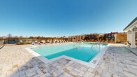 Located at 5315 Waters Edge Village Drive in Charlotte, NC, this outdoor amenity space is a standout feature of the Waters Edge Village community. At its centerpiece is a generously sized, resort-style swimming pool with crystal-clear turquoise water, surrounded by a broad stone-paver deck finished in a classic travertine style. The pool deck is lined with an array of chaise lounges, providing ample space for sunbathing and relaxation under the open Carolina sky.

Adjacent to the pool is a charming covered cabana structure with white columns and decorative lantern lighting, offering shaded seating and a welcoming retreat from the sun. A separate outdoor lounge patio features wicker furniture, a round drum coffee table, and lush landscaping, creating an intimate gathering space ideal for socializing. Multi-level covered balconies on the adjoining clubhouse building overlook the entire pool area, accented with ceiling fans for comfort.

Architectural details throughout the space reflect a refined, coastal-inspired aesthetic with clean white facades, arched windows, and latticed pergola structures that add visual interest and privacy. The surrounding community buildings frame the space, while open sightlines to the sky and mature treeline give the area a bright, airy ambiance throughout the day.