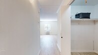 This single-floor unit offers an efficient and thoughtfully arranged layout that maximizes its compact footprint. The combined kitchen and living area forms the heart of the space, featuring light wood-style flooring that runs throughout and bright white walls that enhance the sense of openness. A window with blinds allows natural light to filter into the living area, complementing the wall-unit HVAC system positioned beneath it. Crown molding adds a clean, polished finish to the living space.

The kitchen is a standout feature, equipped with navy blue cabinetry, stainless steel appliances—including a refrigerator, range, and over-the-range microwave—and a peninsula with a stone-finish countertop that provides both additional workspace and a casual dining option. The kitchen flows naturally into the living area, creating a cohesive, open feel.

A dedicated closet with built-in shelving provides organized storage. The bathroom is fully appointed with a tub-shower combination, a dark-vanity with a white countertop and sink, and a large mirror. The overall aesthetic is clean, modern, and neutral, making for a bright and functional living environment.