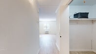 This single-floor unit offers an efficient and thoughtfully arranged layout that maximizes its compact footprint. The combined kitchen and living area forms the heart of the space, featuring light wood-style flooring that runs throughout and bright white walls that enhance the sense of openness. A window with blinds allows natural light to filter into the living area, complementing the wall-unit HVAC system positioned beneath it. Crown molding adds a clean, polished finish to the living space.

The kitchen is a standout feature, equipped with navy blue cabinetry, stainless steel appliances—including a refrigerator, range, and over-the-range microwave—and a peninsula with a stone-finish countertop that provides both additional workspace and a casual dining option. The kitchen flows naturally into the living area, creating a cohesive, open feel.

A dedicated closet with built-in shelving provides organized storage. The bathroom is fully appointed with a tub-shower combination, a dark-vanity with a white countertop and sink, and a large mirror. The overall aesthetic is clean, modern, and neutral, making for a bright and functional living environment.