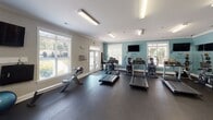 Located at 2041 Berkley Hall Way in Fort Mill, South Carolina, this fitness center offers a well-equipped, single-floor exercise facility designed to support a full range of workout routines. The space is divided into distinct functional zones, each thoughtfully appointed to maximize usability.

The cardio area is bright and welcoming, featuring multiple treadmills, an elliptical trainer, and a rowing machine arranged across durable dark rubber flooring. Large windows with blinds line the walls, flooding the room with natural light and providing views of the surrounding greenery. A wall-mounted flat-screen television adds entertainment for cardio sessions, while French doors offer direct access to the outdoors.

Adjacent to the cardio zone, a dedicated strength training area houses a multi-station cable machine, a dumbbell rack with a full selection of free weights, a weight bench, and additional resistance equipment. Mirrored walls expand the sense of space and allow for proper form checks during workouts. An additional room provides space for functional fitness equipment, including stability balls and exercise mats.

Throughout the facility, crisp white crown molding and soft teal accent walls create a polished, motivating atmosphere. Overhead fluorescent lighting ensures the entire space is well-lit at all times. The overall design balances practicality with an inviting aesthetic, making this fitness center a comprehensive amenity for residents.