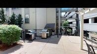 This outdoor communal terrace is a well-appointed amenity space designed for relaxed gatherings and al fresco cooking. The area is anchored by multiple stainless steel gas grills, providing ample grilling capacity for residents and guests alike. The concrete patio surface is spacious and easy to maintain, offering plenty of room to move around the cooking stations comfortably. A covered utility enclosure sits centrally, adding a functional architectural element to the space. The terrace is bordered by sleek black metal railings that define the boundaries of the area while maintaining an open, airy feel. Surrounding multi-story residential buildings frame the space, and mature trees visible beyond the railings contribute a touch of greenery to the urban setting. Additional balconies and walkways of the neighboring structures are visible, reflecting the communal nature of the broader complex. The space benefits from abundant natural light, making it an inviting outdoor destination during daylight hours. Overall, this terrace offers a practical and social outdoor environment well-suited for barbecues, casual gatherings, and enjoying the outdoors within a residential community setting.
