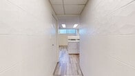 Located at 1 Chapel St in Nanaimo, British Columbia, this single-floor laundry facility offers a clean, functional, and efficiently organized space. The room features light wood-look tile flooring throughout, which complements the crisp white walls and contributes to a bright, hygienic atmosphere. A suspended drop-tile ceiling fitted with recessed panel lighting ensures consistent, even illumination across the entire space. A row of high, horizontal windows along one wall allows natural light to filter in, further enhancing the airy feel of the interior. The facility is equipped with multiple commercial-grade top-loading and front-loading washing machines alongside matching dryers, all neatly arranged to maximize usability. A stainless steel utility sink with a gooseneck faucet provides a practical washing station, complemented by an adjacent stainless steel work surface for handling laundry. White cabinetry offers additional storage and organization. The space also includes a coin-operated payment terminal, a fire extinguisher, and a smoke detector, reflecting attention to safety and operational practicality. Two interior doors lead to additional utility or storage areas. The overall design is straightforward and purposeful, making this a well-appointed shared laundry facility suited to residential or commercial building use.