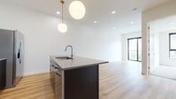 This contemporary single-floor unit showcases a thoughtfully designed open-concept layout that seamlessly connects the kitchen and living area. The kitchen is a standout feature, anchored by a large dark-cabinetry island with an integrated sink and a quartz-style countertop, illuminated by two elegant globe pendant lights suspended from brass fixtures. Stainless steel appliances, including a gas range, microwave, and refrigerator, are complemented by a striking hexagonal tile backsplash and a sleek range hood, all set against warm light oak hardwood flooring that flows throughout the main living space. Recessed lighting and a large window at the far end of the living area invite abundant natural light, creating a bright and airy ambiance. The bedroom features plush carpeting, clean white walls, and a generously sized picture window framing sweeping urban and mountain views. The full bathroom is finished with large-format dark slate tile flooring, a dark-cabinetry vanity with a grey countertop, a framed mirror, and a combination tub and shower, maintaining the cohesive modern aesthetic found throughout the unit.