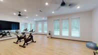 Located at 3125 Fire Ct in Charlotte, NC, this single-level fitness studio is thoughtfully designed to support a range of workout disciplines. The space is divided into distinct functional zones, seamlessly connected by consistent light hardwood-style flooring that runs throughout, lending a clean and cohesive aesthetic to the entire facility.

The primary exercise area is anchored by multiple Cascade cycling bikes arranged to face a large wall-mounted flat-screen display, ideal for guided cycling sessions. A full-length mirror spans one wall, allowing users to monitor their form, while ceiling fans overhead ensure consistent air circulation during intense workouts. Multiple windows dressed with white blinds line the walls, flooding the space with natural light and offering views of the surrounding greenery.

A secondary area provides space for functional and group fitness training. This zone is equipped with a comprehensive rack of fitness accessories, including medicine balls, stability balls of varying sizes, resistance bands, and exercise mats — catering to a wide range of training styles. Framed artwork adds a refined touch to the otherwise utilitarian space.

Additional doors off this area suggest access to locker rooms or storage facilities. Recessed lighting throughout ensures bright, even illumination, while the overall white-walled interior maintains a bright, motivating atmosphere well-suited to an active fitness environment.