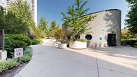Located at 800 S Wells St in Chicago's South Loop neighborhood, this thoughtfully designed outdoor courtyard offers a serene urban retreat within a modern residential complex. The grounds are characterized by a network of gently curving concrete pathways that wind through a richly landscaped environment, guiding visitors through a variety of natural settings.

Mature trees with broad canopies provide generous shade throughout the space, complemented by well-maintained shrubs, ornamental grasses, and layered garden beds filled with diverse plantings. Open lawn areas offer a sense of spaciousness, bordered by dense greenery and defined by sleek black iron fencing that frames the perimeter while maintaining visual connectivity with the surrounding cityscape.

A standout architectural feature is a curved, light-toned building structure at the heart of the courtyard, flanked by large circular white planters housing young trees and ground cover. Circular porthole-style windows accent the structure's facade, adding a distinctive design element. Slim, modern bollard lighting is positioned along the pathways, ensuring the space is functional and inviting at all hours.

The overall ambiance balances urban architecture with natural tranquility, creating a green sanctuary surrounded by the multi-story residential buildings of the complex. The space is well-suited for leisurely strolls, relaxation, and enjoying the outdoors in the heart of Chicago.