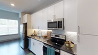 The kitchen is thoughtfully designed with a clean, contemporary aesthetic that balances functionality and style. Crisp white flat-front cabinetry lines the walls from counter to ceiling, maximizing vertical storage while maintaining a bright, open feel. The upper cabinets are fitted with under-cabinet lighting, casting a warm glow across the dark stone countertops below.

A textured hexagonal tile backsplash in white serves as a subtle yet eye-catching focal point, adding dimension to the space without overwhelming its modern palette. The stainless steel appliance suite includes a refrigerator, electric range with ceramic cooktop, over-the-range microwave, and dishwasher—offering a cohesive and polished look throughout.

The single-basin undermount sink is positioned along the central run of cabinetry, paired with a contemporary arc faucet. Dark hardwood-style flooring runs throughout, providing a grounding contrast to the lighter upper elements of the kitchen. The overall design creates an efficient galley-style layout that makes excellent use of the available space, offering ample prep area and storage for everyday living.