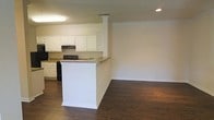 This single-story space showcases an open-concept design that seamlessly connects the kitchen and living area, creating a cohesive and functional layout. The kitchen is outfitted with raised-panel white cabinetry that extends to the upper walls, providing ample storage. A black range and complementary appliances anchor the cooking space, while neutral-toned countertops offer generous workspace. A peninsula with a decorative column detail serves as a natural dividing element between the kitchen and the adjoining living area, making it ideal for casual dining or meal prep. Throughout the space, rich dark hardwood flooring adds warmth and contrast against the light-colored walls. Crown molding runs along the ceiling perimeter, lending a refined, traditional character to the interior. Recessed lighting in the living area and a fluorescent fixture over the kitchen ensure well-distributed illumination. The open layout and neutral color palette create a versatile backdrop suited to a variety of furnishing styles, while the overall design balances practicality with classic detailing.