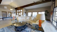 This clubhouse offers a thoughtfully designed communal space that blends contemporary style with warm, inviting elements. The main lounge area is anchored by a striking curved cream sectional sofa, complemented by accent chairs upholstered in geometric patterned fabric, and a bold sculptural coffee table with a dark metal chain-link base and glass top. An area rug in rich, layered tones grounds the seating arrangement, adding depth and texture to the room.

Architectural character is established through an exposed dark wood ceiling beam that spans the width of the space, contrasting beautifully against the light gray walls and vaulted ceiling. Recessed lighting supplements the generous natural light that pours in through multiple windows trimmed in warm wood, offering views of the surrounding exterior. The wood window framing echoes the ceiling beam, creating a cohesive design language throughout.

Decorative accents, including a honeycomb-style metal wall sculpture, glass and metallic side tables, and curated décor objects, add personality and a modern edge to the space. A secondary seating area is visible in the background, extending the usability of the clubhouse. Overall, the space delivers a polished yet comfortable atmosphere well-suited for social gatherings and relaxation.