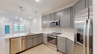 Located at 55 Gardner Dr on Hilton Head Island, South Carolina, this single-floor residence offers a clean, modern design throughout. The kitchen is a standout feature, equipped with gray shaker-style cabinetry, white quartz countertops, a subway tile backsplash, and a full suite of stainless steel appliances including a range, microwave, dishwasher, and refrigerator. Pendant lighting accents the space, while light hardwood-style flooring adds warmth and continuity into the open living area. The living space is bright and airy, benefiting from natural light through large windows and recessed lighting overhead. Built-in shelving adds a practical architectural touch. Bedrooms feature plush carpeting, ceiling fans, and windows that invite natural light, with one bedroom accompanied by a spacious walk-in closet outfitted with wire shelving for ample storage. The bathroom presents a double-sink vanity with white countertops, a large mirror, and a glass-enclosed shower, finished with light tile flooring for a clean, contemporary look. A dedicated laundry area houses a full-size front-loading washer and dryer. The overall ambiance is fresh, bright, and move-in ready, reflecting modern residential design suited for comfortable island living.