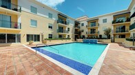 This residential community showcases a striking modern architectural design, characterized by a warm cream and tan exterior facade accented with dark metal balcony railings. The multi-story building wraps around a central courtyard, creating an intimate yet open communal atmosphere bathed in natural sunlight.

At the heart of the outdoor space is a generously sized swimming pool with vivid turquoise water, complemented by a mosaic tile border and a classic red brick paver deck that surrounds it. A water feature adds an elegant touch to the pool area, enhancing the resort-like ambiance of the courtyard.

Each residential level features private balconies with sleek black iron railings, offering residents outdoor space and views of the pool and surrounding landscape. Large windows throughout the building allow for abundant natural light. The ground level is thoughtfully landscaped with manicured greenery and planters that soften the architectural lines and add a welcoming touch to the environment.

The overall design balances a contemporary aesthetic with comfortable, livable details, creating a community space that is both functional and visually appealing. The open-air courtyard layout promotes a sense of community while maintaining a serene and polished atmosphere under the bright open sky.