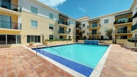 This residential community showcases a striking modern architectural design, characterized by a warm cream and tan exterior facade accented with dark metal balcony railings. The multi-story building wraps around a central courtyard, creating an intimate yet open communal atmosphere bathed in natural sunlight.

At the heart of the outdoor space is a generously sized swimming pool with vivid turquoise water, complemented by a mosaic tile border and a classic red brick paver deck that surrounds it. A water feature adds an elegant touch to the pool area, enhancing the resort-like ambiance of the courtyard.

Each residential level features private balconies with sleek black iron railings, offering residents outdoor space and views of the pool and surrounding landscape. Large windows throughout the building allow for abundant natural light. The ground level is thoughtfully landscaped with manicured greenery and planters that soften the architectural lines and add a welcoming touch to the environment.

The overall design balances a contemporary aesthetic with comfortable, livable details, creating a community space that is both functional and visually appealing. The open-air courtyard layout promotes a sense of community while maintaining a serene and polished atmosphere under the bright open sky.