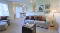 This single-floor residence offers a welcoming and well-connected layout that combines the living, dining, and kitchen areas into one cohesive open-plan space. The living area is comfortably appointed with plush wall-to-wall carpeting and features a large sofa with accent pillows, centered around a dark coffee table. Framed artwork adorns the walls, adding a curated, homey touch to the space. A ceiling fan overhead provides both comfort and subtle visual interest. Natural light flows generously into the space through large sliding glass doors dressed with vertical blinds, creating a bright and airy atmosphere throughout. The dining area sits adjacent to the living room, accommodating a small table and chairs that are ideal for everyday meals. A half-wall partition gracefully separates the kitchen from the main living and dining areas while maintaining the open feel of the floor plan. The kitchen features light wood cabinetry and a pass-through counter that doubles as a breakfast bar, offering both functionality and a sense of connection to the rest of the space. Overall, the layout is practical and inviting, making efficient use of the available space while maintaining a comfortable and livable ambiance.