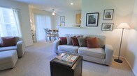 This single-floor residence offers a welcoming and well-connected layout that combines the living, dining, and kitchen areas into one cohesive open-plan space. The living area is comfortably appointed with plush wall-to-wall carpeting and features a large sofa with accent pillows, centered around a dark coffee table. Framed artwork adorns the walls, adding a curated, homey touch to the space. A ceiling fan overhead provides both comfort and subtle visual interest. Natural light flows generously into the space through large sliding glass doors dressed with vertical blinds, creating a bright and airy atmosphere throughout. The dining area sits adjacent to the living room, accommodating a small table and chairs that are ideal for everyday meals. A half-wall partition gracefully separates the kitchen from the main living and dining areas while maintaining the open feel of the floor plan. The kitchen features light wood cabinetry and a pass-through counter that doubles as a breakfast bar, offering both functionality and a sense of connection to the rest of the space. Overall, the layout is practical and inviting, making efficient use of the available space while maintaining a comfortable and livable ambiance.