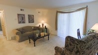 This single-floor living space features a well-appointed sitting area designed for comfort and functionality. The living room is anchored by a plush sofa adorned with decorative pillows, paired with a patterned accent chair that adds visual interest. A dark wood coffee table serves as the centerpiece, styled with decorative objects that complement the room's neutral color palette. Wall art featuring classic urn motifs adds a traditional touch to the space.

A large floor-to-ceiling window dressed with vertical blinds and a warm-toned valance allows abundant natural light to fill the room while offering privacy control. The light beige carpeting runs throughout, contributing to the warm and inviting ambiance. A table lamp provides soft ambient lighting, enhancing the room's cozy atmosphere during evening hours.

The ceiling features a ventilation grille, indicating a central HVAC system for year-round comfort. The overall design blends traditional furnishings with neutral tones, creating a cohesive and relaxed environment suited for everyday living and entertaining.