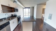 Located on the ninth floor at 624 Yale Avenue in Seattle's South Lake Union neighborhood, this modern unit offers a thoughtfully designed open-concept layout across a single floor. The kitchen is a standout feature, equipped with stainless steel appliances including an electric range and over-the-range microwave, sleek white cabinetry, and a dark-topped island that provides generous prep and dining space. The kitchen flows seamlessly into the living area, creating an expansive, connected environment ideal for both everyday living and entertaining. Exposed ductwork and a concrete ceiling lend an industrial character to the space, complementing the rich, dark hardwood flooring that runs throughout. Floor-to-ceiling windows at the far end of the living area flood the interior with natural light and frame sweeping views of the surrounding cityscape and water beyond — a defining feature of this elevated Seattle address. The neutral concrete and white tones of the walls and ceilings create a clean, contemporary backdrop, while the open sightlines enhance the overall sense of space and airiness. Additional utility areas are accessible off the main living space, rounding out the functional layout of this well-appointed urban residence.