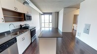 Located on the ninth floor at 624 Yale Avenue in Seattle's South Lake Union neighborhood, this modern unit offers a thoughtfully designed open-concept layout across a single floor. The kitchen is a standout feature, equipped with stainless steel appliances including an electric range and over-the-range microwave, sleek white cabinetry, and a dark-topped island that provides generous prep and dining space. The kitchen flows seamlessly into the living area, creating an expansive, connected environment ideal for both everyday living and entertaining. Exposed ductwork and a concrete ceiling lend an industrial character to the space, complementing the rich, dark hardwood flooring that runs throughout. Floor-to-ceiling windows at the far end of the living area flood the interior with natural light and frame sweeping views of the surrounding cityscape and water beyond — a defining feature of this elevated Seattle address. The neutral concrete and white tones of the walls and ceilings create a clean, contemporary backdrop, while the open sightlines enhance the overall sense of space and airiness. Additional utility areas are accessible off the main living space, rounding out the functional layout of this well-appointed urban residence.