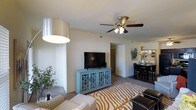 This single-floor residence offers a thoughtfully designed open-concept layout that connects the living, dining, and kitchen areas into one cohesive and functional living environment. The living area features plush wall-to-wall carpeting, creating a warm and comfortable atmosphere, and is anchored by a ceiling fan with integrated lighting that provides both air circulation and ambient illumination. Neutral-toned walls throughout the space lend a clean, versatile backdrop that complements a variety of décor styles. A smooth transition leads from the living area into the dining space, which accommodates a full dining set and benefits from natural light filtering in from nearby windows. The kitchen is accessible directly from the dining area, reinforcing the open and connected feel of the floor plan. A hallway visible from the main living space provides access to additional rooms, suggesting a well-organized and practical layout. The space makes efficient use of its footprint, balancing openness with defined zones for relaxing, dining, and entertaining. Overall, the residence presents a bright, airy ambiance with a comfortable and livable character suitable for everyday living.