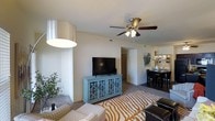 This single-floor residence offers a thoughtfully designed open-concept layout that connects the living, dining, and kitchen areas into one cohesive and functional living environment. The living area features plush wall-to-wall carpeting, creating a warm and comfortable atmosphere, and is anchored by a ceiling fan with integrated lighting that provides both air circulation and ambient illumination. Neutral-toned walls throughout the space lend a clean, versatile backdrop that complements a variety of décor styles. A smooth transition leads from the living area into the dining space, which accommodates a full dining set and benefits from natural light filtering in from nearby windows. The kitchen is accessible directly from the dining area, reinforcing the open and connected feel of the floor plan. A hallway visible from the main living space provides access to additional rooms, suggesting a well-organized and practical layout. The space makes efficient use of its footprint, balancing openness with defined zones for relaxing, dining, and entertaining. Overall, the residence presents a bright, airy ambiance with a comfortable and livable character suitable for everyday living.