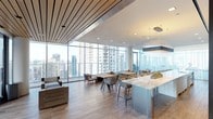 This refined amenity lounge occupies a high floor of a contemporary urban high-rise, offering an impressive and well-appointed space designed for both relaxation and social gatherings. The centerpiece of the room is an expansive wall of floor-to-ceiling glass windows that frames breathtaking panoramic views of the surrounding city skyline, flooding the interior with abundant natural light throughout the day.

The space is thoughtfully divided into distinct zones. A communal dining area features a large wooden table surrounded by woven, high-back chairs, ideal for group gatherings or shared meals. Adjacent to this is a striking kitchen island finished in rich, veined marble with a light gray base, complemented by a dramatic crystal pendant chandelier overhead that adds a touch of glamour to the space.

A comfortable lounge seating area with plush sofas and accent chairs provides a more relaxed setting near the windows, perfect for taking in the urban vistas. The flooring throughout consists of wide-plank wood-look tiles in warm neutral tones, lending warmth and continuity to the open-plan layout.

Overhead, a slatted wood ceiling feature adds architectural interest and texture, transitioning elegantly into smooth white recessed-lit ceilings. The overall ambiance is one of polished, contemporary sophistication, balancing form and function in a bright, airy setting.