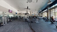 This expansive fitness center is thoughtfully designed to accommodate a full range of workout needs within a single, open-plan floor. The facility is accessed through a welcoming entry lobby featuring soaring ceilings, modern pendant globe lighting, dark-framed glass walls, and a built-in wooden cubby storage wall — offering a clean and organized transition into the workout space.

The main fitness floor is divided into distinct training zones. The strength training area is lined with floor-to-ceiling mirrors and equipped with a comprehensive selection of selectorized cable machines, multi-use weight stations, dumbbell racks with adjustable benches, and additional resistance equipment. Battle ropes are also available for functional training. Multiple wall-mounted flat-screen TVs are positioned throughout for entertainment.

The cardio zone runs along the perimeter, bathed in natural light from expansive floor-to-ceiling windows that overlook the surrounding outdoor landscape. Treadmills, ellipticals, stair climbers, and stationary bikes are neatly arranged in rows, each offering views to the exterior.

Overhead, vaulted ceilings with pendant industrial-style lights and ceiling fans create an airy, comfortable atmosphere throughout. The flooring consists of durable, dark-toned carpet tile, tying the modern aesthetic together. The overall ambiance is bright, clean, and professional, suited for a high-traffic fitness environment.