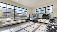 This contemporary fitness facility is thoughtfully designed to support a wide range of workout styles within a single, well-appointed level. The space is defined by soaring ceilings and expansive floor-to-ceiling windows framed in sleek black steel, flooding the interior with abundant natural light and offering open views of the surrounding outdoor amenity area. Light wood-style flooring runs throughout, complementing the clean, neutral color palette of the white walls and trim.

The main training area is generously proportioned, featuring full-length mirrored walls that enhance the sense of space and support form-focused exercise. Plyo boxes are positioned throughout for functional and plyometric training. A dedicated equipment zone houses an extensive rack system stocked with a full range of kettlebells, medicine balls, and resistance accessories, providing comprehensive strength and conditioning options.

A separate spin studio features multiple stationary cycling bikes and is decorated with motivational "Positive Vibes" wall art, creating an energetic and encouraging atmosphere. Mirrored walls and additional natural light from corner windows round out this focused training room.

The facility also includes a connecting corridor with a sanitation station, reinforcing a clean and well-maintained environment. The overall design is modern, bright, and purpose-built for an active lifestyle.