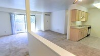 This single-floor residence offers a clean, open-concept layout that connects the main living area to the kitchen in a seamless, functional design. The living space is finished with wall-to-wall carpeting that extends throughout, lending a comfortable and cohesive feel to the interior. Crisp white walls keep the space feeling airy and bright, while a large sliding glass door along one wall invites generous natural light and provides views of the lush greenery outside. The open sightlines created by the half-wall divider enhance the sense of spaciousness, making the area feel expansive and connected. Overhead HVAC vents and a baseboard heating unit ensure year-round climate comfort. The adjacent kitchen features light wood cabinetry and a neutral countertop surface, offering a practical and tidy cooking space that flows naturally into the main living area. Structural column accents and clean architectural lines give the interior a simple, understated character. The overall ambiance is light, neutral, and move-in ready, providing a versatile canvas well-suited for a variety of living arrangements and interior styles.
