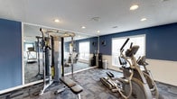 This fitness center offers a thoughtfully designed workout environment suited for a variety of exercise routines. The space features bold navy blue accent walls complemented by crisp white wainscoting, creating a modern and energizing atmosphere. Recessed lighting throughout the ceiling, combined with large windows that allow ample natural light, ensures the space remains bright and welcoming at all times. The textured ceiling adds a classic finish, while the patterned carpet flooring provides comfort underfoot during workouts. A full cable machine system with a functional trainer serves as a central strength-training station, complete with an adjustable bench for a wide range of exercises. Cardio equipment, including an elliptical trainer, is positioned to take advantage of the natural light from the windows. A large floor-to-ceiling mirror along one wall visually expands the space and allows users to monitor their form during exercise. A water station is conveniently available within the room. The overall layout is open and organized, offering a clean, functional, and motivating environment for residents or members to pursue their fitness goals.