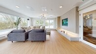 Located at 5828 Granville Street in Vancouver, BC, this thoughtfully designed single-floor space offers a bright, open-plan layout that seamlessly blends comfort and functionality. The interior is unified by warm light wood flooring throughout, crisp white walls, and a clean contemporary aesthetic.

The living lounge area anchors one end of the space, featuring generous sectional seating, ottomans, and a large wall-mounted TV set into a light wood feature panel. A long, backlit low-profile media console adds a refined touch, while multiple wide windows flood the area with natural light and offer views of the surrounding greenery.

Toward the center, a games area with a shuffleboard table provides an additional recreational zone, connected by glass-paneled doors to an adjacent corridor. The kitchen is well-appointed with light wood cabinetry, a bold dark countertop with a deep blue backsplash, stainless steel appliances, open shelving, and a farmhouse-style sink — combining practicality with modern style.

The dining area is generously sized, accommodating a long communal table and multiple chairs, with floor-to-ceiling sliding glass doors and large windows opening to outdoor views, filling the room with an airy, welcoming atmosphere. The overall ambiance is modern, calm, and community-oriented.