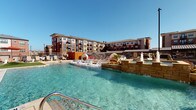 This outdoor pool area is designed as a comprehensive resort-style retreat, set against the backdrop of a multi-story brick building with balconied upper floors. The centerpiece is a generously sized swimming pool with crystal-clear water, featuring built-in in-water lounge chairs that allow residents to relax partially submerged while enjoying open-air views and abundant natural sunlight.

The surrounding concrete deck is thoughtfully arranged with multiple distinct activity zones. A standout fire pit seating area incorporates a sleek linear gas fire feature with blue glass accents, flanked by striped cushioned sofas and wicker lounge chairs, ideal for evening gatherings. String lights suspended overhead add a warm ambiance to the patio after dark.

An outdoor kitchen station, constructed with stone masonry and stainless steel appliances, includes a full-sized gas grill, prep surface, sink, and storage, all sheltered beneath a pergola-style overhead structure. Nearby, a long communal dining table provides ample space for al fresco meals. Additional seating vignettes are scattered throughout the courtyard, offering flexible social spaces. The entire area is enclosed by iron fencing, ensuring privacy while maintaining an open, airy atmosphere.