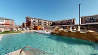 This outdoor pool area is designed as a comprehensive resort-style retreat, set against the backdrop of a multi-story brick building with balconied upper floors. The centerpiece is a generously sized swimming pool with crystal-clear water, featuring built-in in-water lounge chairs that allow residents to relax partially submerged while enjoying open-air views and abundant natural sunlight.

The surrounding concrete deck is thoughtfully arranged with multiple distinct activity zones. A standout fire pit seating area incorporates a sleek linear gas fire feature with blue glass accents, flanked by striped cushioned sofas and wicker lounge chairs, ideal for evening gatherings. String lights suspended overhead add a warm ambiance to the patio after dark.

An outdoor kitchen station, constructed with stone masonry and stainless steel appliances, includes a full-sized gas grill, prep surface, sink, and storage, all sheltered beneath a pergola-style overhead structure. Nearby, a long communal dining table provides ample space for al fresco meals. Additional seating vignettes are scattered throughout the courtyard, offering flexible social spaces. The entire area is enclosed by iron fencing, ensuring privacy while maintaining an open, airy atmosphere.