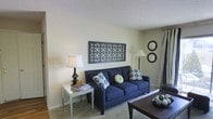 This single-floor space offers a warm and inviting atmosphere centered around a thoughtfully arranged living area. The room is finished with neutral, light-toned walls that create a bright and airy feel, complemented by carpeted flooring that adds comfort underfoot. A textured ceiling adds a subtle architectural detail overhead.

The living area serves as the heart of the space, anchored by a deep navy blue sofa adorned with decorative patterned throw pillows in blue, green, and yellow tones. A dark wood coffee table sits at the center, providing both function and style. A mirrored side table beside the sofa holds a classic table lamp, adding warm ambient lighting to the room.

Wall décor enhances the space's character, with a large geometric lattice-style wall panel serving as a striking focal point above the sofa, while a pair of round circular mirrors nearby adds visual depth and balance. A floor lamp positioned in the corner further supplements the lighting.

Curtain panels along the wall suggest a window or access point that allows for natural light. The overall ambiance is cozy and well-curated, blending classic furnishings with decorative accents to create a lived-in yet stylish environment suitable for relaxing and entertaining.