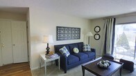 This single-floor space offers a warm and inviting atmosphere centered around a thoughtfully arranged living area. The room is finished with neutral, light-toned walls that create a bright and airy feel, complemented by carpeted flooring that adds comfort underfoot. A textured ceiling adds a subtle architectural detail overhead.

The living area serves as the heart of the space, anchored by a deep navy blue sofa adorned with decorative patterned throw pillows in blue, green, and yellow tones. A dark wood coffee table sits at the center, providing both function and style. A mirrored side table beside the sofa holds a classic table lamp, adding warm ambient lighting to the room.

Wall décor enhances the space's character, with a large geometric lattice-style wall panel serving as a striking focal point above the sofa, while a pair of round circular mirrors nearby adds visual depth and balance. A floor lamp positioned in the corner further supplements the lighting.

Curtain panels along the wall suggest a window or access point that allows for natural light. The overall ambiance is cozy and well-curated, blending classic furnishings with decorative accents to create a lived-in yet stylish environment suitable for relaxing and entertaining.