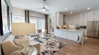 This single-floor residence offers a thoughtfully designed open-concept layout that seamlessly connects the living, dining, and kitchen areas. The living room is bright and welcoming, featuring warm wood-look flooring, a ceiling fan, recessed lighting, and large sliding glass doors with decorative patterned drapes that invite natural light and provide outdoor views. The kitchen is a standout feature, showcasing crisp white shaker-style cabinetry extending to the ceiling, a light quartz peninsula countertop with an undermount sink, a subway tile backsplash with under-cabinet lighting, a wall-mounted range hood, and stainless steel appliances including a French door refrigerator. The bedroom is generously proportioned with plush carpeting, a ceiling fan, a bold accent wall, large windows with outdoor views, and direct access to the en-suite bathroom. The primary bathroom features a dual-sink vanity with white shaker cabinetry, quartz countertops, and wide mirrors with overhead lighting. A second bathroom offers a deep soaking tub and shower combination with large-format gray tile surround, wood-look flooring, and a tall white storage cabinet. Throughout the residence, a cohesive neutral palette with contemporary accents creates a polished and comfortable ambiance.