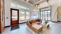 Located at 28266 Huntsville Brownsferry Rd in Madison, Alabama, this thoughtfully designed single-floor space offers a compelling blend of contemporary and industrial aesthetics. The layout encompasses an open living area and a kitchenette, creating a versatile, flowing environment suited for gathering and entertaining.

The living area is immediately striking, anchored by soaring ceilings with decorative coffered detailing accented in warm copper tones. A bold, multi-drum pendant chandelier in black and gold serves as a central focal point. Floor-to-ceiling windows along the front façade, supplemented by transom windows above, flood the interior with abundant natural light and offer clear views of the surrounding streetscape. A full-panel glass garage-style door further blurs the boundary between interior and exterior. A sleek linear fireplace set into large-format tile anchors one wall, above which hangs a statement abstract artwork.

An adjacent lounge area features deep navy accent walls with geometric metal detailing, tiled flooring with an intricate star pattern, and casual high-top seating. The compact kitchenette showcases ornate patterned tile, a slate-toned countertop, mixed-finish cabinetry, and stainless steel appliances. Throughout the space, durable large-format tile and hardwood-style flooring complement the refined, modern-industrial ambiance.