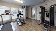This single-floor fitness center is thoughtfully designed to provide a comprehensive workout experience in a clean, functional environment. The space is finished with durable hardwood-style flooring throughout, lending a warm and polished aesthetic that complements the neutral, light-toned walls.

The cardio area is equipped with a True-brand treadmill and a True elliptical machine, both positioned near large windows fitted with horizontal blinds that allow ample natural light to fill the space. A wall-mounted television is conveniently located near the entry, providing entertainment during workouts.

The strength training section features a multi-station cable machine, a full rack of dumbbells ranging across multiple weight increments, and an adjustable workout bench. A wall-mounted exercise mat and a posted workout chart provide additional support for varied training routines. A large floor-length mirror is mounted on the wall, helping users monitor their form and visually expanding the sense of space.

Fluorescent overhead lighting ensures the interior remains well-lit at all times. The fitness center also includes a dedicated entry door marked with an illuminated exit sign, restroom access indicated by signage, and a fire safety device on the wall. The overall ambiance is practical and inviting, catering to a range of fitness levels and workout styles.