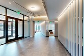 Located at 2 West 4th Street in downtown Wilmington, Delaware, this thoughtfully designed amenity floor offers a sophisticated blend of modern architecture and functional spaces. The layout spans a single generously sized floor, encompassing two hallways, an exercise room, and a living room.

The hallways make an immediate architectural statement, featuring striking vertical slat wall panels in light wood and white tones that extend from floor to ceiling and continue as ceiling accents with integrated ambient lighting. Large-format dark stone-look tile flooring grounds the space, while recessed lighting and a sculptural pendant fixture at the entrance add visual interest. Floor-to-ceiling glass entry doors invite abundant natural light and offer views of the surrounding streetscape.

The living room is defined by a curved wall of vertical slats that filters soft natural light from the exterior, creating a warm, lounge-like atmosphere. A plush curved sectional sofa and complementary furnishings make this an inviting retreat.

The exercise room is bright and well-appointed, with large windows overlooking the street, providing natural light and outdoor views. It features cardio equipment including treadmills, ellipticals, and cycling bikes, along with free weights, a weight bench, and mirrored walls. Carpet tile flooring and a built-in water fountain complete the functional amenity space.