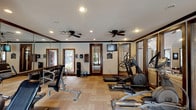 This fitness center offers a thoughtfully designed workout environment that balances functionality with an inviting aesthetic. The space features warm wood crown molding and door trim that complement the light parquet wood flooring, creating a refined atmosphere uncommon in typical gym settings. Multiple ceiling fans are installed throughout, ensuring consistent air circulation during workouts, while recessed lighting provides bright, even illumination across the entire floor.

The cardio zone is well-appointed with multiple treadmills and elliptical trainers positioned to allow comfortable spacing between users. A wall-mounted flat-screen television is conveniently placed for entertainment during cardio sessions. The free weight area includes a fully stocked dumbbell rack with a wide range of weights to accommodate various fitness levels, accompanied by a water cooler station for easy hydration.

Full-length mirrored wall panels line one side of the room, visually expanding the space and providing users with proper form feedback during exercise. Large windows with wood-framed surrounds allow natural light to filter in and offer views of the surrounding outdoor greenery. A glass partition wall connects the fitness center to an adjacent lounge or common area, maintaining an open, airy feel throughout. The overall ambiance is clean, comfortable, and purpose-built for effective training.
