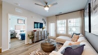 This single-story residence offers a thoughtfully designed open-concept layout finished with light wood-style flooring that flows consistently throughout the space. The living room serves as the heart of the unit, featuring a ceiling fan with integrated lighting, neutral walls, and large windows with an exterior door that invite abundant natural light and outdoor views. A wall-mounted television and rustic media console anchor the room, while the adjacent accent wall adds a contemporary touch.

The kitchen is well-appointed with white shaker-style cabinetry extending to the ceiling, stainless steel appliances including a refrigerator, range, microwave, and dishwasher, and grey stone countertops. A generously sized island with pendant lighting overhead provides additional prep space and casual seating for multiple guests.

The primary bedroom is a comfortable retreat with large windows framing outdoor greenery, wood-style flooring, and a ceiling fan. Its en-suite bathroom features a long vanity with grey countertops, white cabinetry, a broad mirror, and direct access to a spacious walk-in closet with built-in shelving. A second bedroom mirrors the inviting atmosphere with ample natural light and stylish finishes throughout.