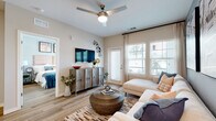 This single-story residence offers a thoughtfully designed open-concept layout finished with light wood-style flooring that flows consistently throughout the space. The living room serves as the heart of the unit, featuring a ceiling fan with integrated lighting, neutral walls, and large windows with an exterior door that invite abundant natural light and outdoor views. A wall-mounted television and rustic media console anchor the room, while the adjacent accent wall adds a contemporary touch.

The kitchen is well-appointed with white shaker-style cabinetry extending to the ceiling, stainless steel appliances including a refrigerator, range, microwave, and dishwasher, and grey stone countertops. A generously sized island with pendant lighting overhead provides additional prep space and casual seating for multiple guests.

The primary bedroom is a comfortable retreat with large windows framing outdoor greenery, wood-style flooring, and a ceiling fan. Its en-suite bathroom features a long vanity with grey countertops, white cabinetry, a broad mirror, and direct access to a spacious walk-in closet with built-in shelving. A second bedroom mirrors the inviting atmosphere with ample natural light and stylish finishes throughout.