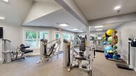 Located at 100 Hunt Club Trail in Marlton, NJ, this fitness center offers a well-equipped, single-floor exercise facility designed to accommodate a wide range of workout needs. The space features a vaulted ceiling in select areas, contributing to an open and airy atmosphere, while flush-mounted panel lighting ensures consistent, bright illumination throughout.

Large windows along multiple walls flood the interior with natural light and provide pleasant views of the surrounding greenery, creating an inviting environment for exercise. Full-length mirrored walls along one side visually expand the space and support proper form during workouts.

The facility is comprehensively outfitted with Matrix-brand cardio equipment, including treadmills, ellipticals, and a stationary cycle, all positioned to take advantage of the outdoor views. The strength training area houses multiple selectorized resistance machines, a dedicated free-weight station with a dumbbell rack, and a variety of functional fitness accessories including stability balls, resistance bands, medicine balls, and foam rollers organized on a dedicated storage rack.

Neutral-toned carpet tiles cover the flooring throughout, offering comfort underfoot. A wall-mounted television, a small reception or resource counter, and wall art add functional and aesthetic touches. The overall ambiance is clean, modern, and purpose-driven, making this fitness center a practical and welcoming space for residents of the Hunt Club Trail community.