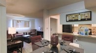 This single-floor residence offers an open-concept design that seamlessly connects the main living areas. The dining area, positioned at the foreground, features a glass-top table surrounded by upholstered chairs in rich burgundy tones, accented by decorative centerpieces that add a touch of elegance. A breakfast bar with light countertops separates the dining space from the kitchen, which is equipped with a black refrigerator and ample counter space, making it both functional and accessible.

The living room beyond is carpeted and furnished with plush seating, including a patterned armchair and additional soft furnishings, creating a comfortable and inviting atmosphere. A window with vertical blinds allows natural light to filter into the living area. Neutral wall tones throughout the space contribute to a cohesive and understated aesthetic.

A doorway visible from the main living area provides access to additional rooms, suggesting a practical and well-organized floor plan. Decorative wall art, including a wine-themed framed print in the kitchen area, adds personality to the space. Overall, the layout is efficient and thoughtfully arranged for everyday living.