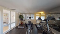 This single-level fitness room offers a dedicated space for cardiovascular exercise, outfitted with multiple treadmills, elliptical trainers, and a stationary bike arranged across durable rubber-tile flooring. The room is finished in neutral tones, with warm sconce lighting complementing the natural light that filters in through several windows fitted with blinds. Full-length mirrors line portions of the walls, visually expanding the space and allowing users to monitor their form during workouts. A wall-mounted flat-screen television provides entertainment options during exercise sessions. Decorative touches include a potted plant that adds a subtle natural element to the otherwise functional environment. The entrance features glass-paneled doors and a framed interior window looking into an adjacent tiled common area, creating a sense of openness and connectivity between spaces. A stool is positioned near the entry, and a branded welcome mat marks the threshold. The overall layout is practical and organized, designed to accommodate multiple users simultaneously while maintaining a clean, uncluttered atmosphere suited for focused exercise.
