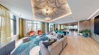 This contemporary amenity lounge is a thoughtfully designed social space that seamlessly blends bold artistic expression with comfortable, functional living. The space is anchored by a striking coffered ceiling adorned with an intricate graphic pattern and warm ambient lighting, creating an immediate focal point upon entry. Light hardwood flooring flows throughout, lending warmth and continuity across the open layout.

The main lounge area features generous sectional seating arranged around a patterned area rug, complemented by vibrant coral accent chairs that inject a lively pop of color. Adjacent to this is a dedicated game area with a round gaming table and matching coral chairs, positioned alongside floor-to-ceiling windows dressed in sheer olive-green curtains that filter natural light while framing expansive urban views.

A wet bar with a mosaic tile backsplash and integrated cabinetry serves as a refined entertaining hub, flanked by a dining table with teal upholstered chairs. A media lounge nook features plush rounded seating, a wall-mounted television, and a distinctive textured accent wall.

The space also includes a slatted wood ceiling section that adds organic texture, along with lush tropical plants that bring the outdoors in. Throughout, the design balances energetic color palettes with sophisticated materials, making this an inviting and versatile communal destination.