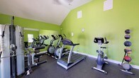 This fitness center at 14510 Lemoyne Blvd in Biloxi, Mississippi, is a well-appointed, single-floor facility designed to support a comprehensive workout experience. The space is immediately recognizable by its bold lime-green walls and striking vaulted ceilings, which lend an open, energetic atmosphere throughout. Durable rubber flooring covers the workout area, providing safety and comfort underfoot.

The facility is equipped with a variety of cardio machines, including treadmills, ellipticals, and stationary bikes, several of which are reflected in a large wall mirror that enhances the sense of space. A multi-station cable and weight machine anchors the strength training section, while a dedicated free weights area features a full rack of dumbbells, exercise mats, and medicine balls on a vertical storage rack.

Natural light filters in through multiple windows fitted with blinds, and a ceiling fan helps maintain comfortable airflow. A wall-mounted flat-screen television adds entertainment options during workouts. French doors open to an outdoor area, further brightening the space and providing easy access to the exterior. The entrance is marked by a branded welcome mat, reinforcing the community feel of the facility. Overall, the gym offers a functional, motivating environment for users of all fitness levels.