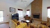Located at 201 Eads Street in East Ridge, Tennessee, this single-story home offers a comfortable and well-appointed living environment. The living room serves as the centerpiece of the space, showcasing soaring vaulted ceilings that create an open, airy atmosphere. A full-height brick fireplace anchors one wall, adding a classic and rustic charm to the room. A ceiling fan is mounted overhead, providing both functionality and a finishing touch to the cathedral ceiling. Natural light filters in through multiple windows dressed with elegant valances and blinds, brightening the neutral-toned walls and carpeted floors. The living area is generously sized, accommodating a plush seating arrangement that includes floral and plaid upholstered furniture centered around a traditional wood coffee table, making it an ideal space for relaxing or entertaining. The warm color palette of golds, greens, and burgundies throughout the furnishings complements the brick and neutral wall tones, lending the home a cozy, welcoming ambiance. Overall, this East Ridge residence combines classic architectural details with comfortable living spaces, creating a warm and inviting home.