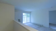 This single-floor space features a clean, minimalist design characterized by neutral white walls and smooth ceilings that contribute to an open, airy atmosphere. The main living area is finished with wall-to-wall carpeting, providing a comfortable and quiet underfoot experience. A prominent sliding glass door serves as the focal point of the room, flooding the interior with abundant natural light and providing direct access to a private balcony. Through the glass, mature trees are visible, offering a pleasant, nature-facing outlook. A baseboard heating unit runs along the exterior wall beneath the sliding door, ensuring efficient climate control throughout the colder months. A standard interior door is also visible, connecting to an adjacent room or closet space. The overall layout is straightforward and functional, with clean lines and uncluttered wall surfaces that allow for flexible furniture arrangement. The bright, evenly lit environment, combined with the outdoor connection provided by the balcony access, creates a welcoming and practical living space suited to a variety of lifestyles.