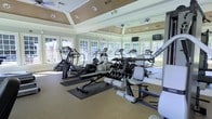 This single-story fitness center offers a well-appointed workout environment designed for both cardio and strength training. The space is framed by large, multi-pane windows on multiple walls, flooding the interior with abundant natural light and providing views of the surrounding greenery outdoors. Full-length mirrors line the walls, visually expanding the room and allowing users to monitor their form during exercise.

The ceiling is a notable architectural feature, showcasing a tray design with warm wood paneling and recessed lighting, lending the space an elevated, upscale character. A ceiling fan is centrally mounted, contributing to air circulation throughout workouts.

The flooring consists of durable, cushioned fitness-grade material well-suited for high-impact activities. Equipment is thoughtfully arranged throughout the space, including elliptical trainers, a rowing machine, and a variety of strength and resistance machines. Exercise accessories such as stability balls and dumbbells are also available. Wall-mounted televisions provide entertainment during workouts. Overall, the space balances functionality with a bright, refined aesthetic.
