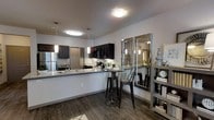 This single-floor residence offers a sleek and modern living environment centered around an open-concept layout that seamlessly connects the kitchen and dining areas. The kitchen is outfitted with rich dark cabinetry, granite-look countertops, and stainless steel appliances, including a full-size refrigerator and built-in microwave. A wide peninsula with pendant lighting overhead serves as both a prep space and casual bar-height seating area, making it ideal for entertaining or everyday dining. The flooring throughout features dark hardwood-style planks that lend warmth and continuity to the space. A compact dining nook is positioned adjacent to the kitchen, complete with a round bistro-style table and industrial bar stools. A decorative open shelving unit and a large framed mirror add depth and visual interest to the dining area, enhancing the sense of openness. Recessed and flush-mount ceiling lighting ensures the space remains bright and well-illuminated. The overall ambiance is contemporary and well-appointed, with a cohesive dark-toned aesthetic balanced by neutral walls that keep the interior feeling fresh and inviting.