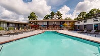 Located at 420 N Dean Rd in Auburn, Alabama, this outdoor amenity area serves as a central gathering space within the community. The focal point is a generously sized swimming pool with classic brick tile edging and crystal-clear blue water, designed to accommodate both relaxation and recreation. The pool is surrounded by a broad, open concrete sundeck that offers ample room for lounging, furnished with rows of modern chaise lounge chairs and accented by bright blue market umbrellas that provide shade throughout the day. On one side of the deck, a red pergola structure serves as a dedicated outdoor lounge and grilling area, complete with wicker seating and a built-in grill — ideal for social gatherings. Stone planter beds and manicured landscaping line the perimeter of the deck, adding natural texture and greenery to the hardscape. The surrounding buildings feature a two-story design with covered walkways and balconies, creating a courtyard-style atmosphere that encloses the space. Towering mature trees frame the background, providing privacy and a lush, natural backdrop. The open sky above allows for abundant natural light, making this outdoor space bright and inviting. The overall design balances functionality with aesthetic appeal, offering a well-appointed outdoor retreat in the heart of Auburn.