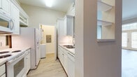 This single-floor residence offers a thoughtfully designed layout with a clean, neutral palette and a variety of functional spaces. The galley-style kitchen is outfitted with white cabinetry extending to the ceiling, a double stainless steel sink, white appliances including a refrigerator with ice dispenser, electric range, and built-in microwave, all set atop light hardwood-style flooring. A dedicated laundry area is conveniently accessible directly from the kitchen. The living area features light hardwood-style flooring, crown molding, and a pass-through opening to the kitchen, complemented by built-in shelving along one wall. The entry area is highlighted by elegant French doors flanked by sidelights and a transom window, flooding the space with natural light. The carpeted bedrooms are generously sized; one features a ceiling fan, large windows with natural views, and ample wall space, while another boasts elevated ceilings, a transom window above the doorway, and access to a spacious walk-in closet. The bathroom presents a long white vanity with an extended countertop, a large mirror, pendant lighting, tile flooring, and a full soaking tub with shower. Throughout the residence, warm beige tones and light finishes create a bright, welcoming ambiance.